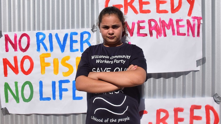 A young girl crosses her arms and stands with attitude infront of a corrugated iron fence with activist signs
