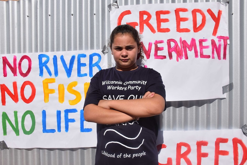 A young girl crosses her arms and stands with attitude infront of a corrugated iron fence with activist signs