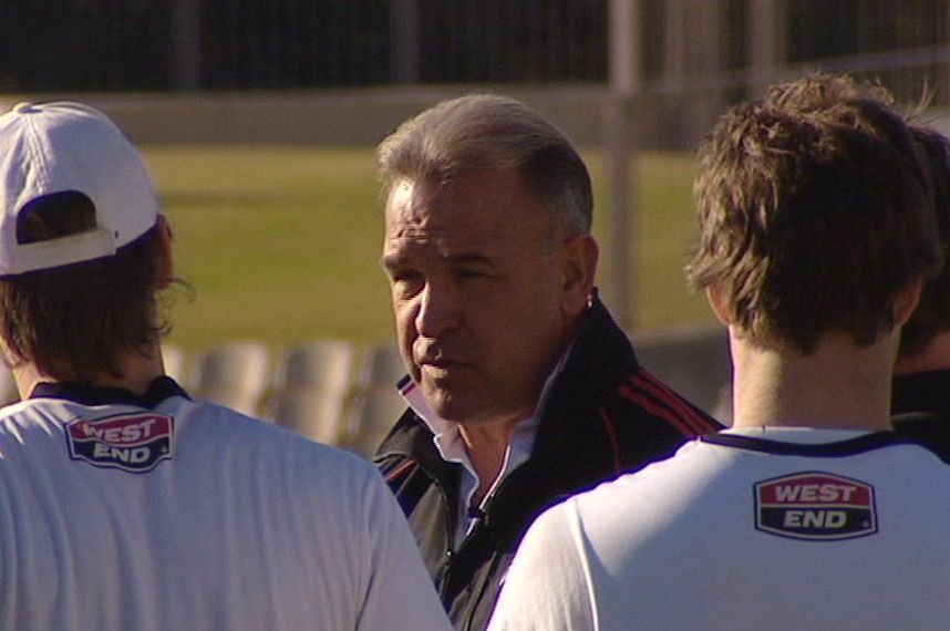 A man wearing a sports coat stands surrounded by players on a cricket field