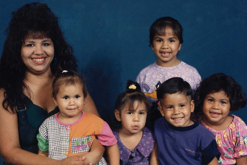 A young woman sits with one toddler on her lap and four other children sitting next to her in a smiley family portrait