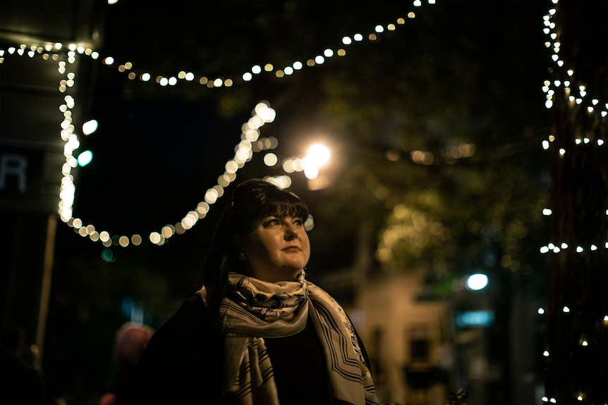 Sherrie D'Souza looks up into the distance, wears scarf and dark jacket and stands on street with fairy lights at night.