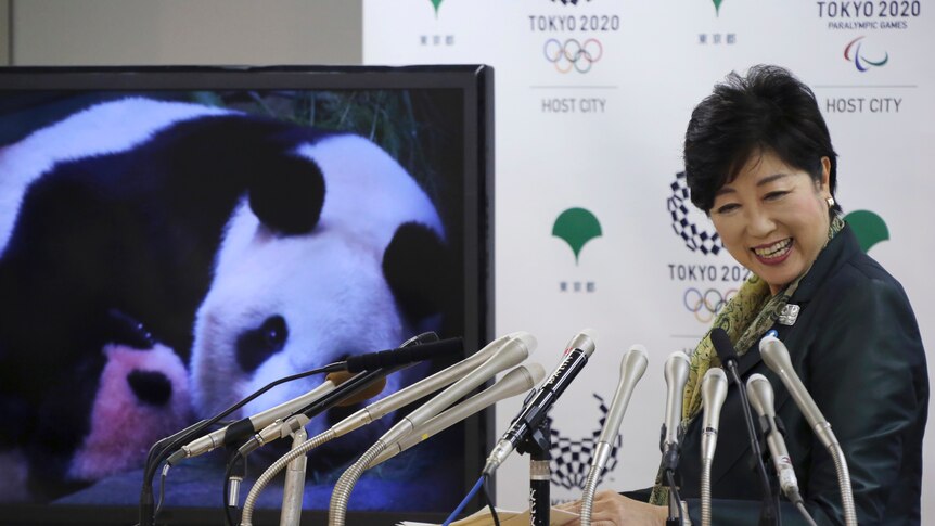Yuriko Koike standing next to a screen shows an image of Japan's female baby giant panda held by her mother.