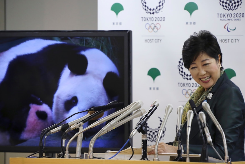 Yuriko Koike standing next to a screen shows an image of Japan's female baby giant panda held by her mother.