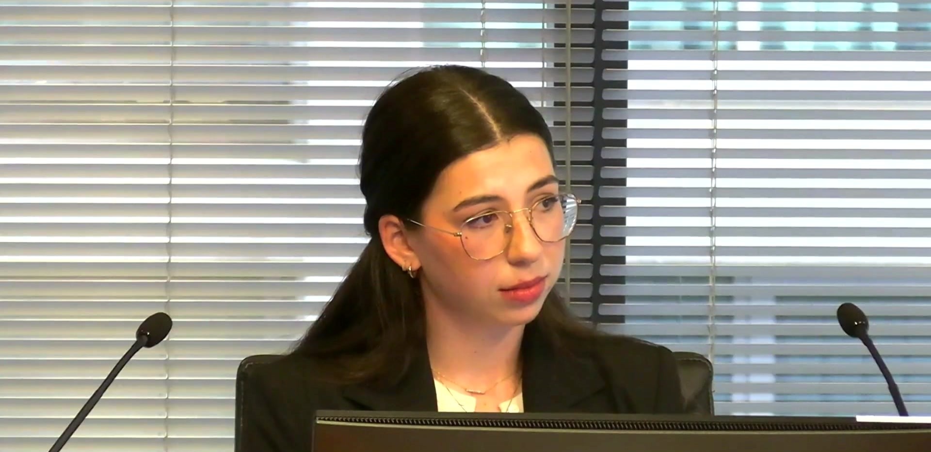 A woman with long dark hair pulled back in a ponytail in a black jacket with clear rimmed glasses sits behind two microphones.
