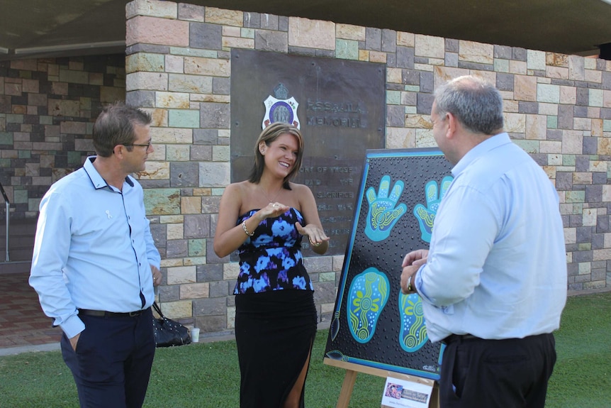 Three people stand discussing a painting placed on an easel