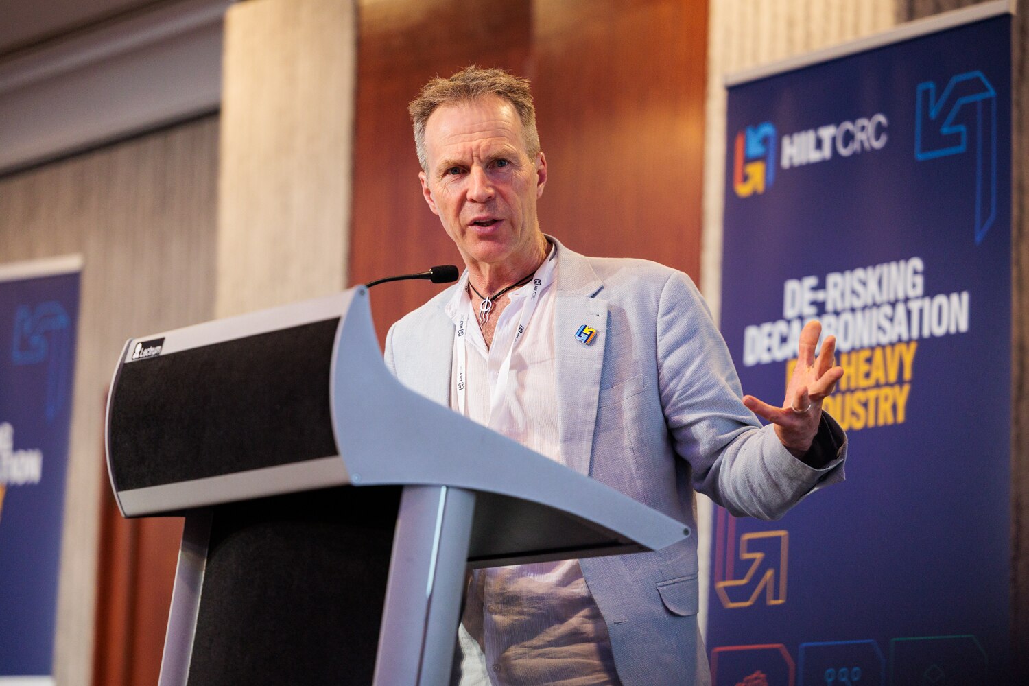Un hombre con traje gris claro y camisa blanca se encuentra detrás de un podio y hace gestos con las manos. 