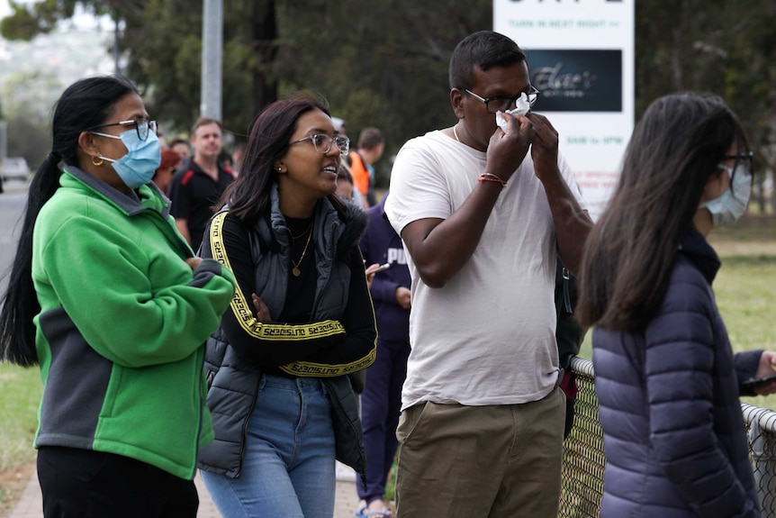 Four young people, two wearing masks and two not, including one man blowing his nose into a tissue.