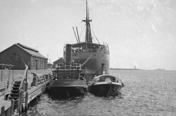 Black and white photo of ship, tug boat and small launch tied up at wooden wharf