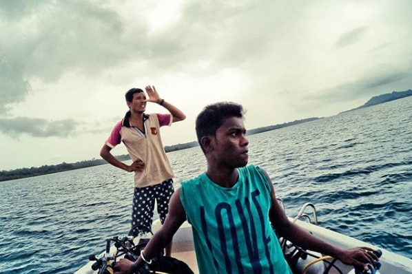 John Allen Chau stands at the rear of a boat while a dark skinned man sits in the front guiding a small boat on the ocean.