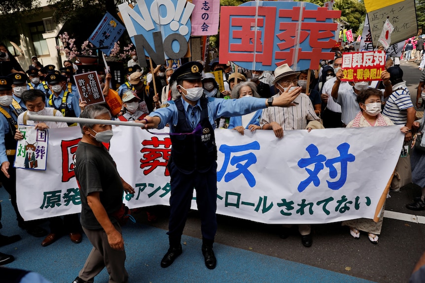 A police officer stands before a large crowd of protesters, who hold signs with Japanese characters.