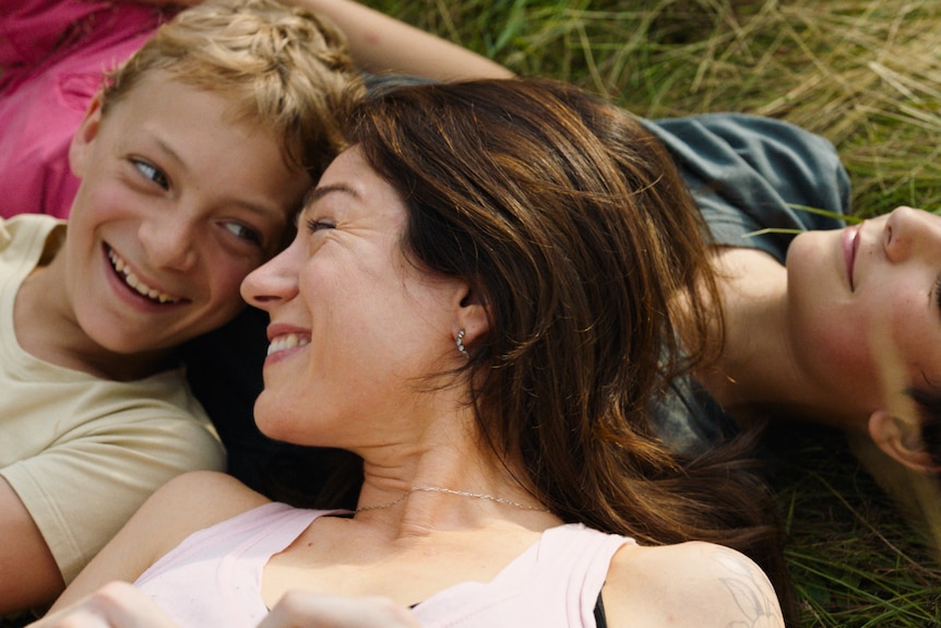 A middle-aged brunette woman lies in the grass, smiling. A teenage boy laughs beside her, another smiling content behind.