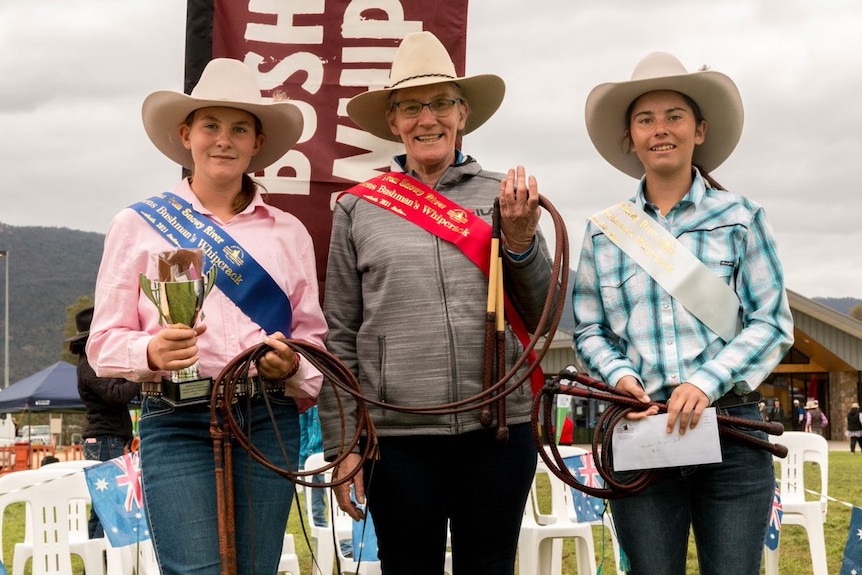 Three women with cowboy hats stand with ribbons across their chests, holding trophies and holding a whip.