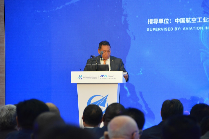 Peter Shanel stands behind a lecturn giving a speech on the conference stage. 