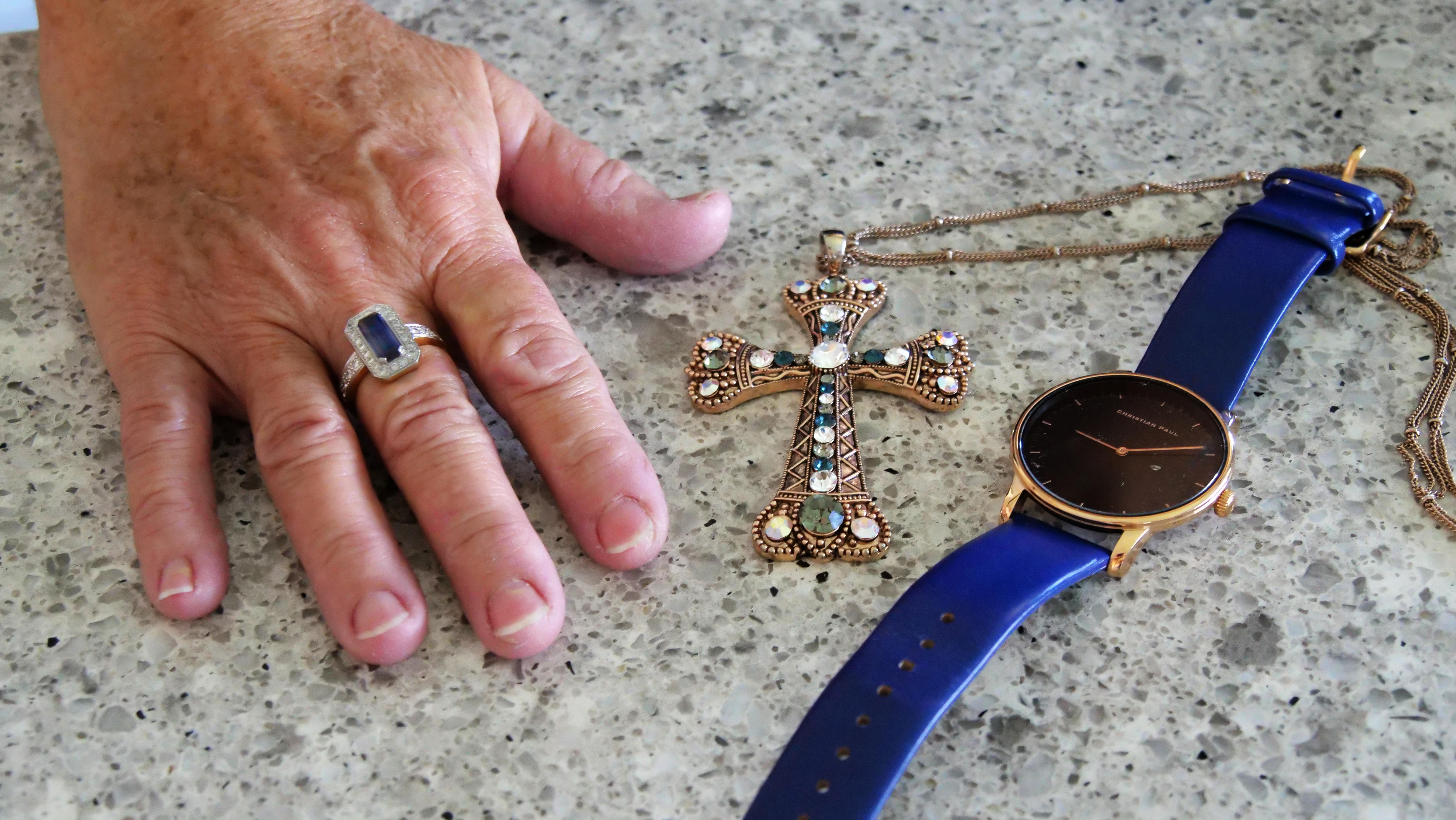 A womans hand with blue ring, next to a golden cross, and watch with blue band and gold face