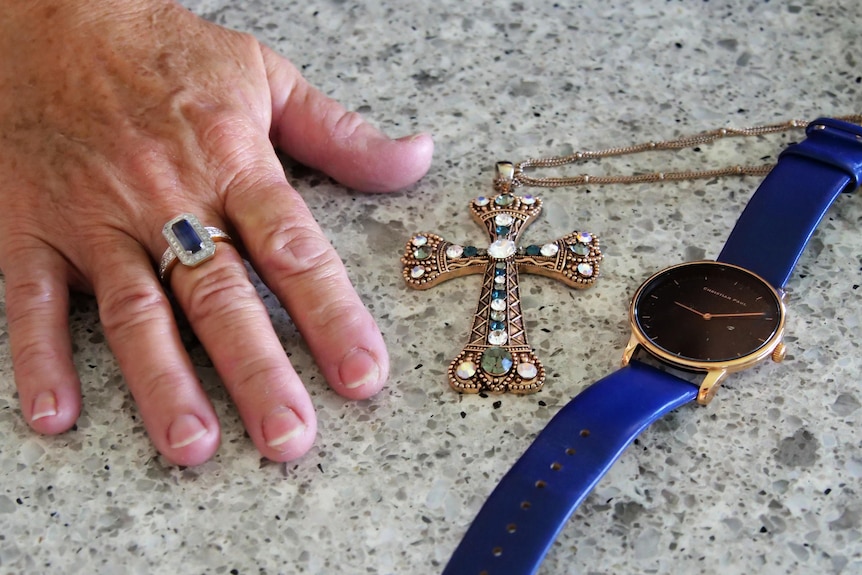 A womans hand with blue ring, next to a golden cross, and watch with blue band and gold face