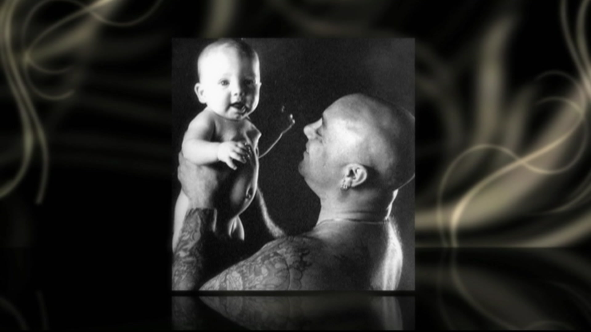 A black and white photo of a bald man with tattooed arms looking at a baby he's holding up while the baby looks at the camera.