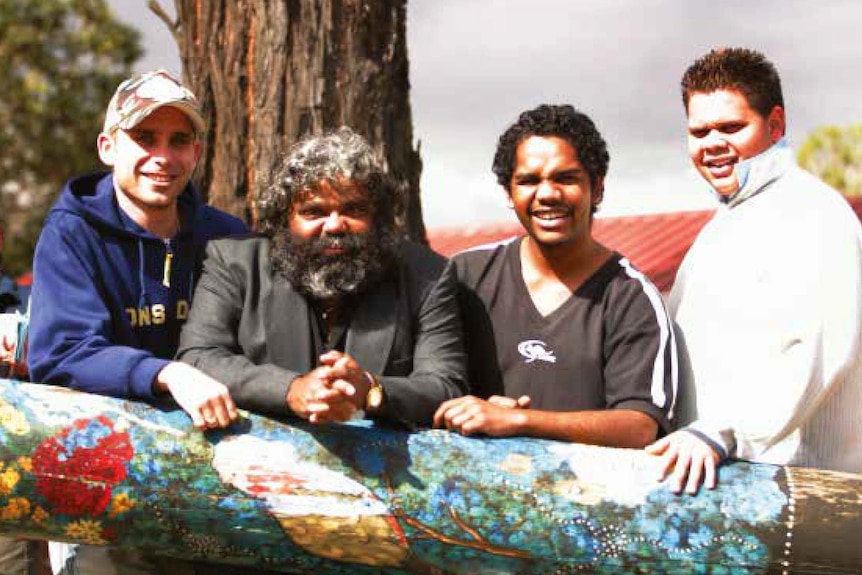 Four Indigenous men stand leaning against a large didgeridoo, smiling and squinting into the sun. 