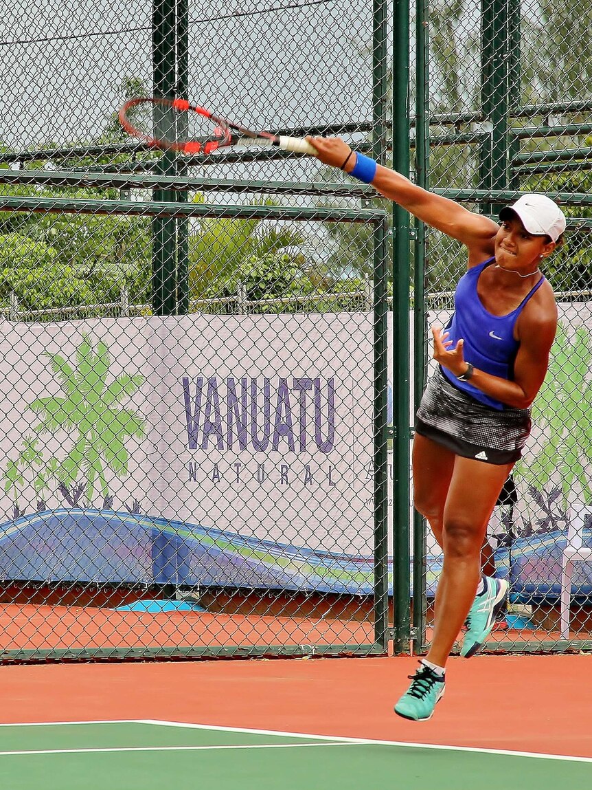 An athletic Samoan woman playing tennis jumps of the ground after smashing an overhead ball.