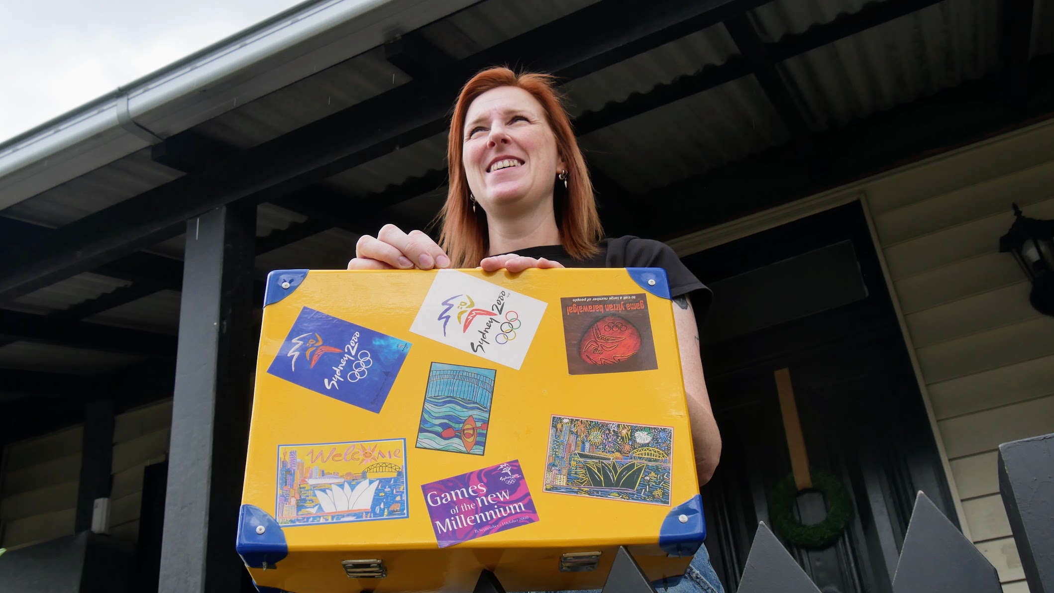 Uma mulher olha para longe e segura uma mala amarela com adesivos das Olimpíadas de Sydney.