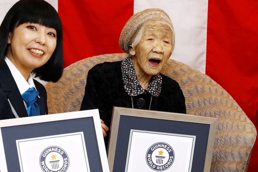 An elderly Japanese woman sits next to a younger Japanese woman, as both smile and hold Guinness World Record certificates.