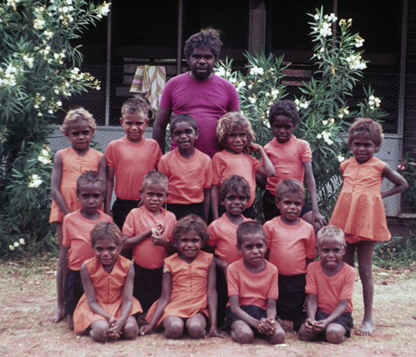 A group of Aboriginal school kids stand in a group. An Aboriginal man stands behind them