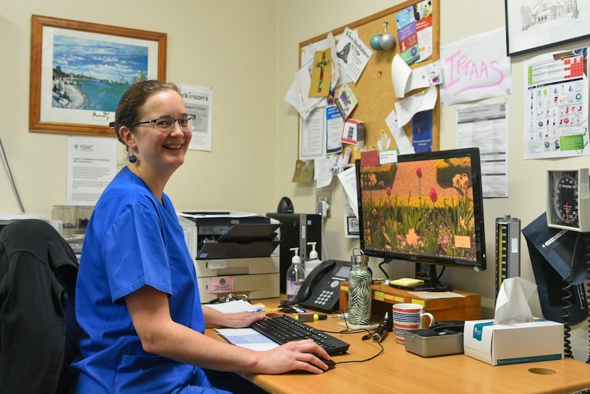 A smiling woman with glasses and blue scrubs is sitting at a desk in a doctor's office.