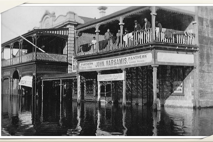 Two old buildings stand in a flood, black and white photo. The building days 'John Varsamis Pantheon Cafe Palace'