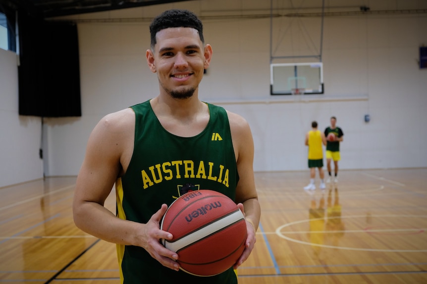 Jarrod wears a green and gold Australia singlet and smiles at the camera, while holding a basketball.