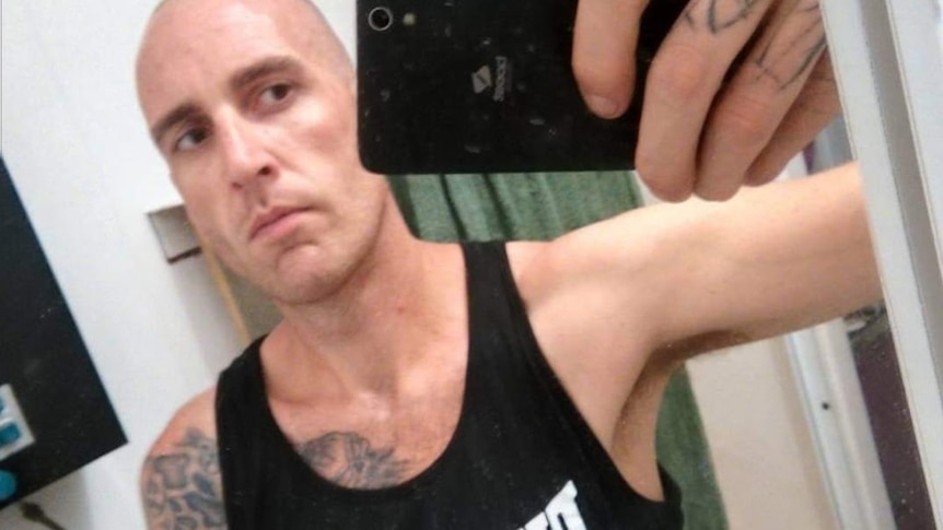 A bald man in a singlet, with tattoos visible on his arm and chest, takes a selfie with a phone.