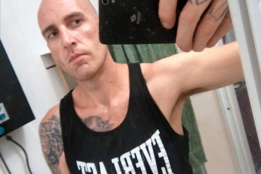 A man in a black singlet with a shaved head holds phone up to take a selfie in mirror.