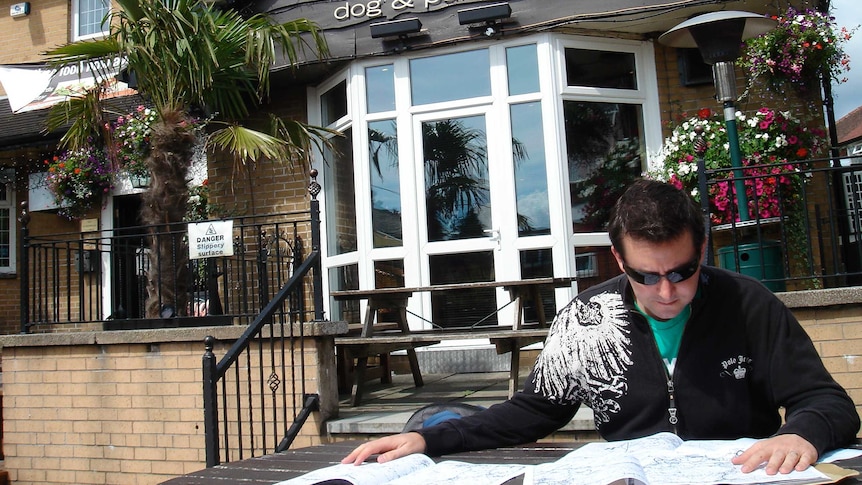 A man in sunglasses sits at a table covered with maps outside the Dog and Partridge weatherboard pub.