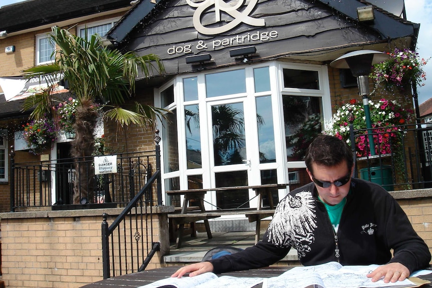 A man in sunglasses sits at a table covered with maps outside the Dog and Partridge weatherboard pub.