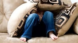 A boy sits on a couch, with his head poking through cushions he is covered with.