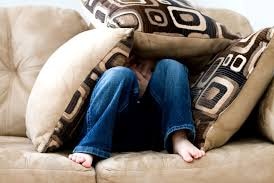 A boy sits on a couch, with his head poking through cushions he is covered with.
