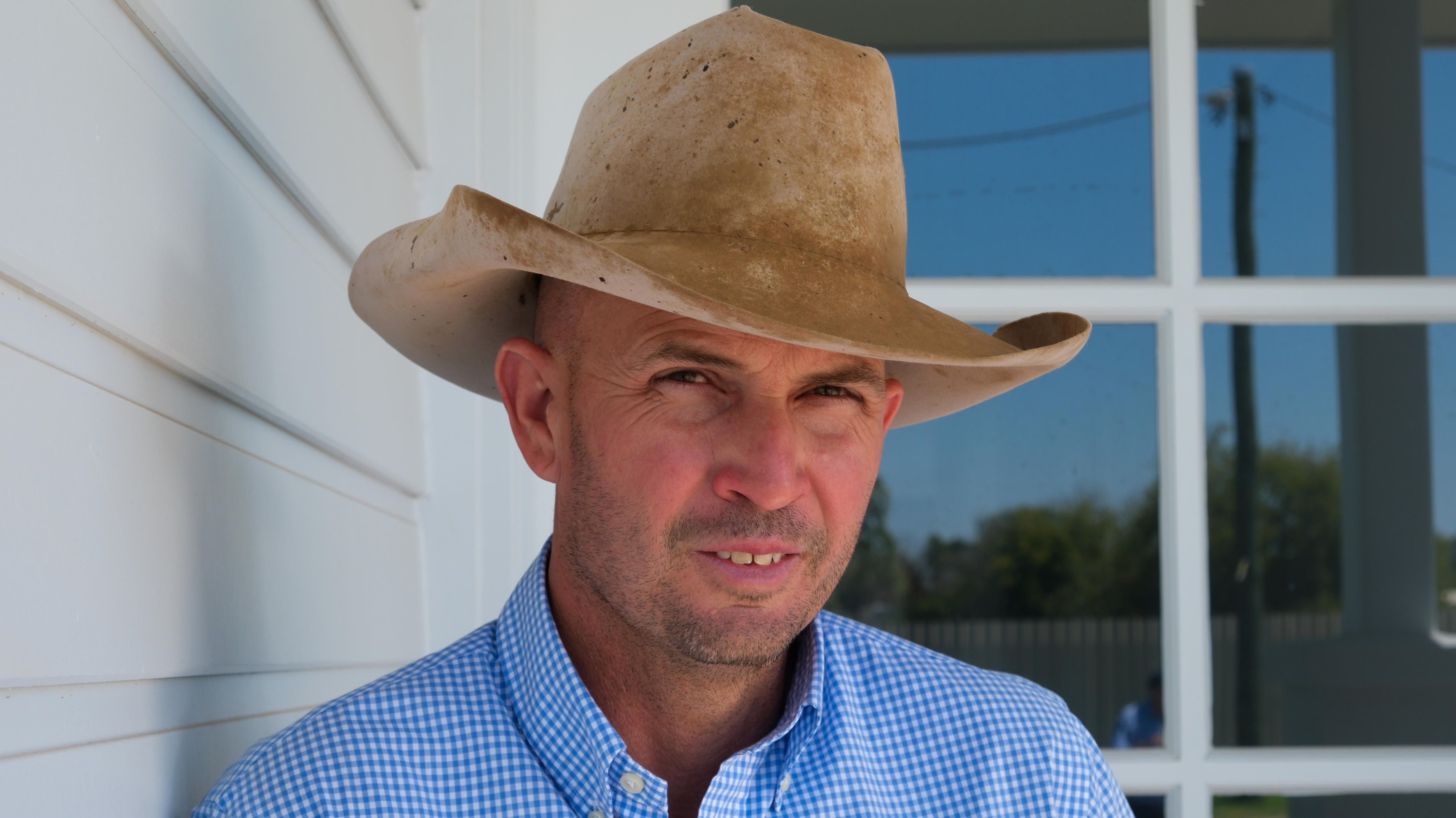 Un hombre con un sombrero de estilo country se apoya contra una pared, mirando hacia adelante.