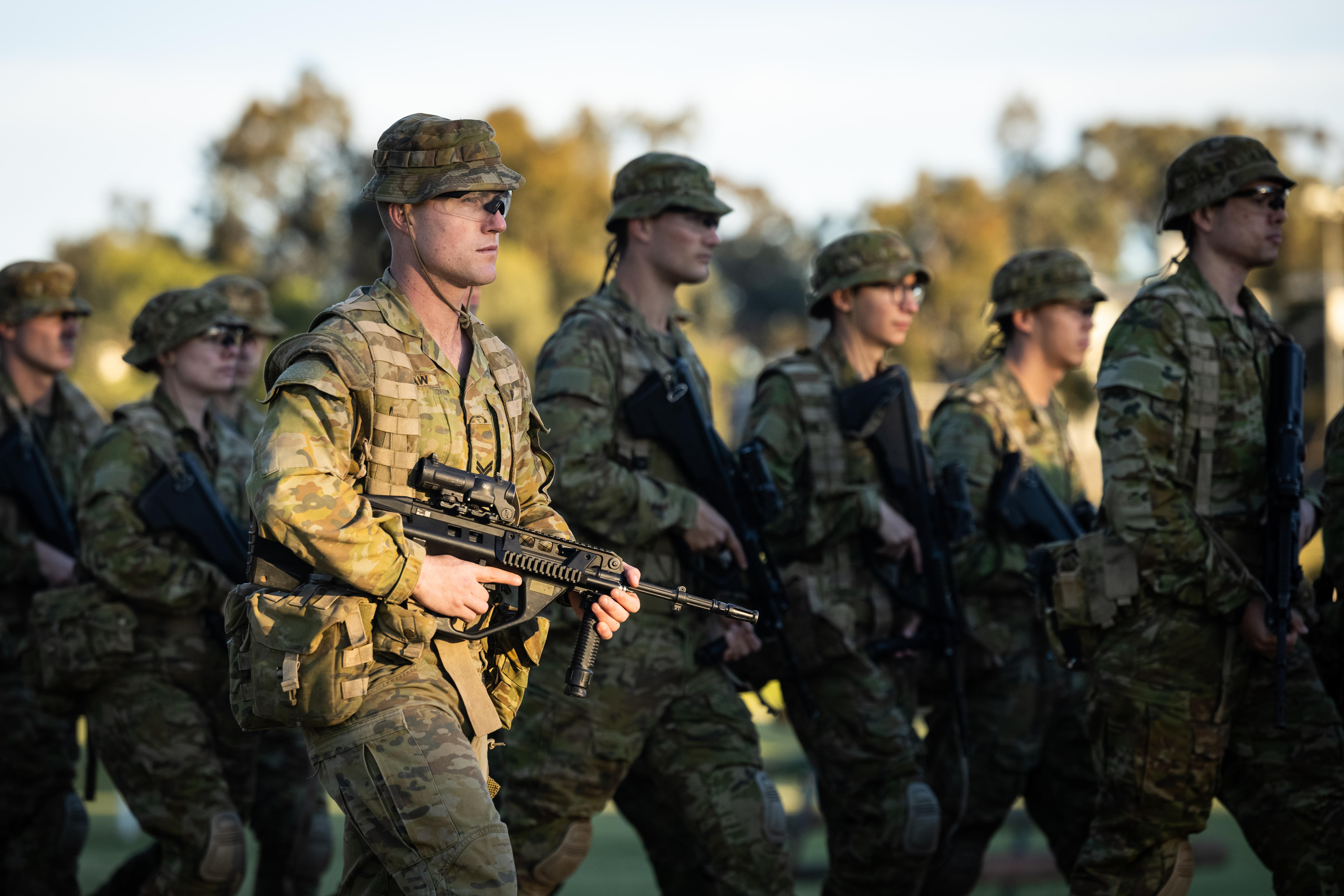 Man wearing army greens, hat and clear glasses walks from left to right, carrying a large black gun.