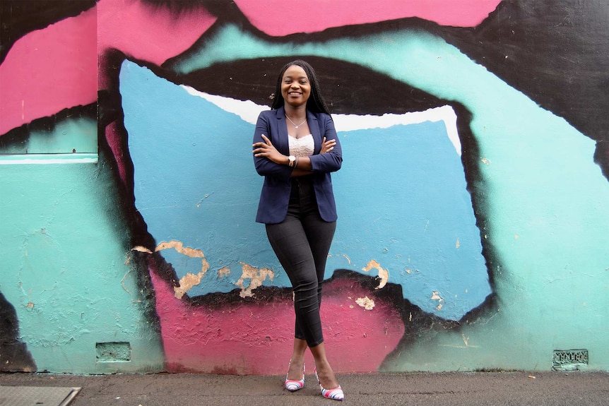 Prudence Melom smiles with her arm crossed, standing in front of a colourful mural in Toowoomba.