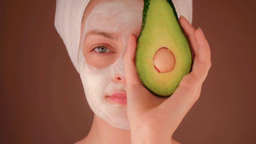 A woman with a beauty face mask holds half an avocado over her eye