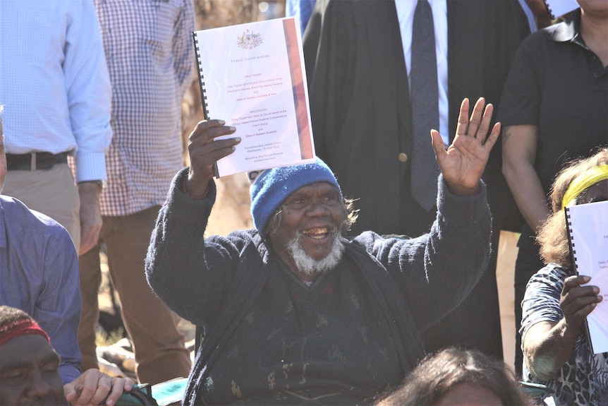 An Indigenous person in a blue beanie holds a document happily above their head