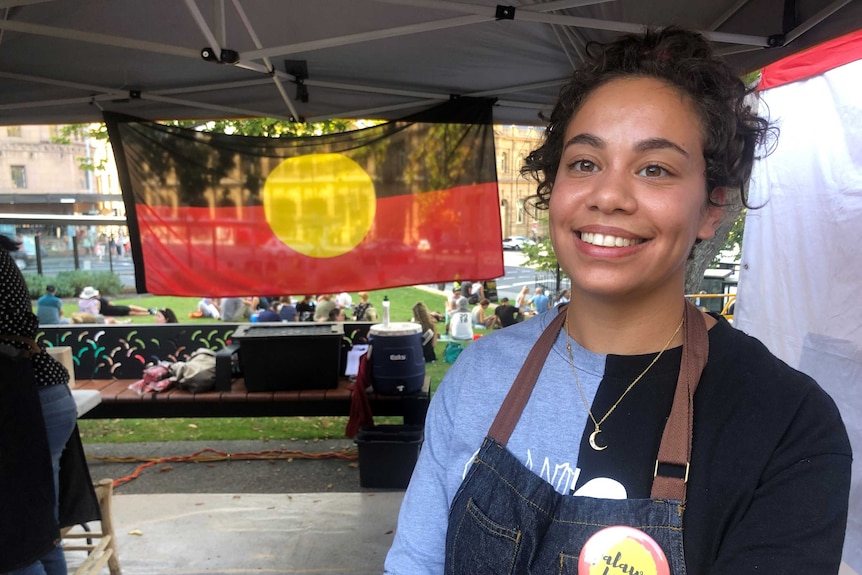 Nunami Sculthorpe stands at food stall smiling. An Aboriginal flag hangs in the background.