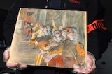 Close view of a small painting showing a line of men singing is held in the gloved hands of an unidentified person