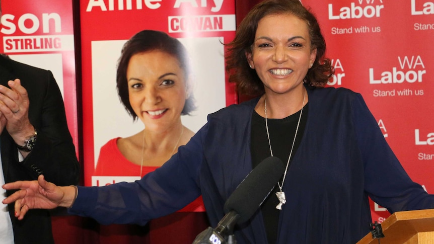A smiling Anne Aly stands on stage in front of a microphone and red Labor signage featuring her face.