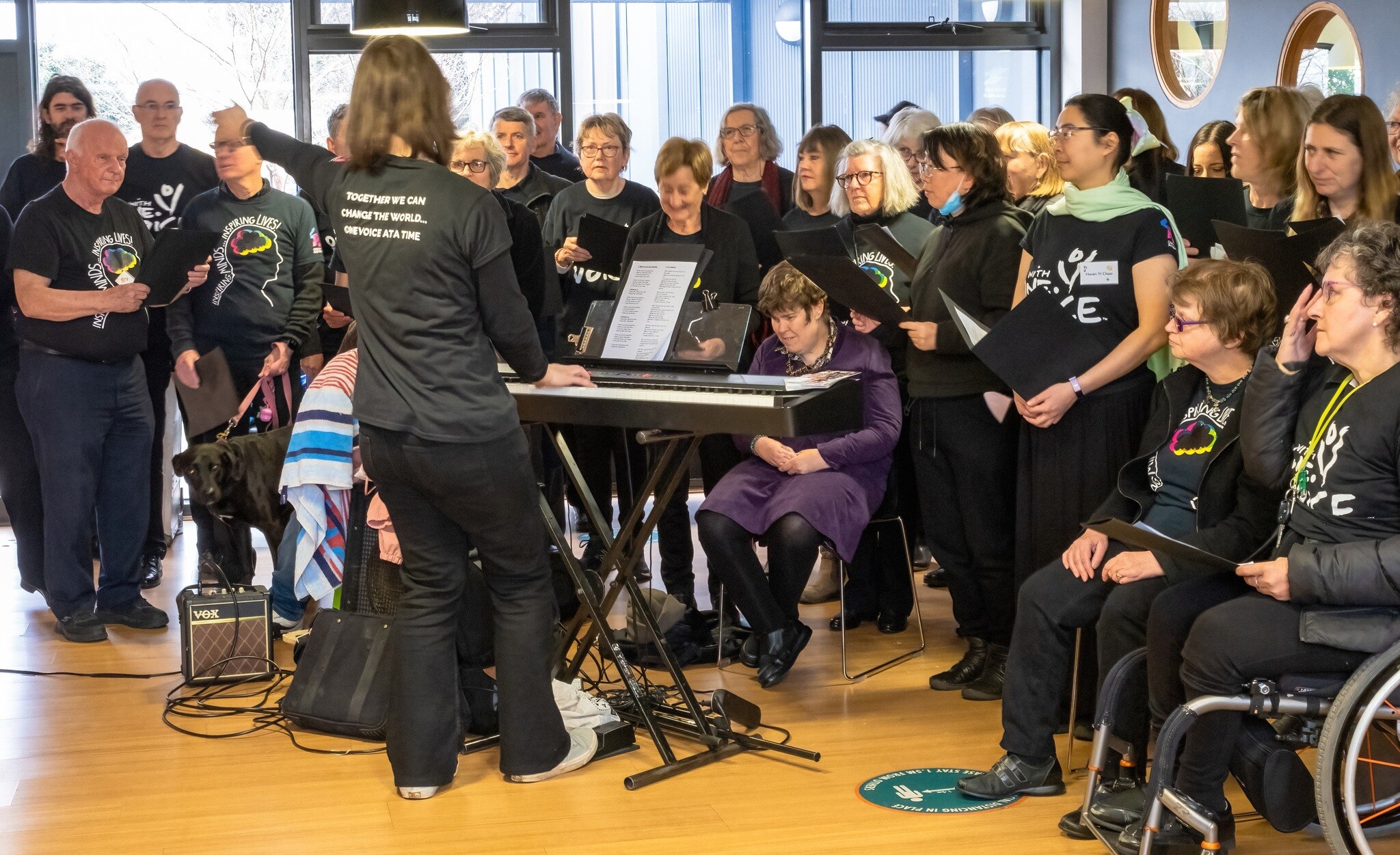 Ashburton With One Voice choir performs in black group tshirts with Kym Dillon playing keyboard and conducting in front of them.