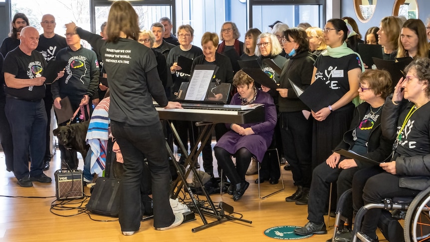 Ashburton With One Voice choir performs in black group tshirts with Kym Dillon playing keyboard and conducting in front of them.