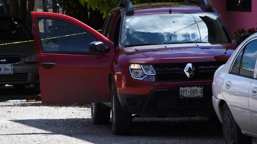 A red car with a bullet hole at the top of the windscreen and police tape in the background.