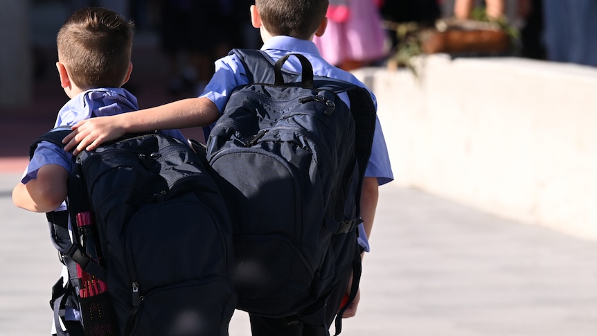 Two young school boys walking with their backpacks on as one hugs the other.