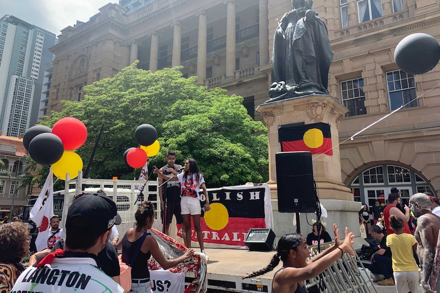 A woman stands and speaks into a microphone with one hand raised. There are black, red and yellow balloons and Indigenous flags.
