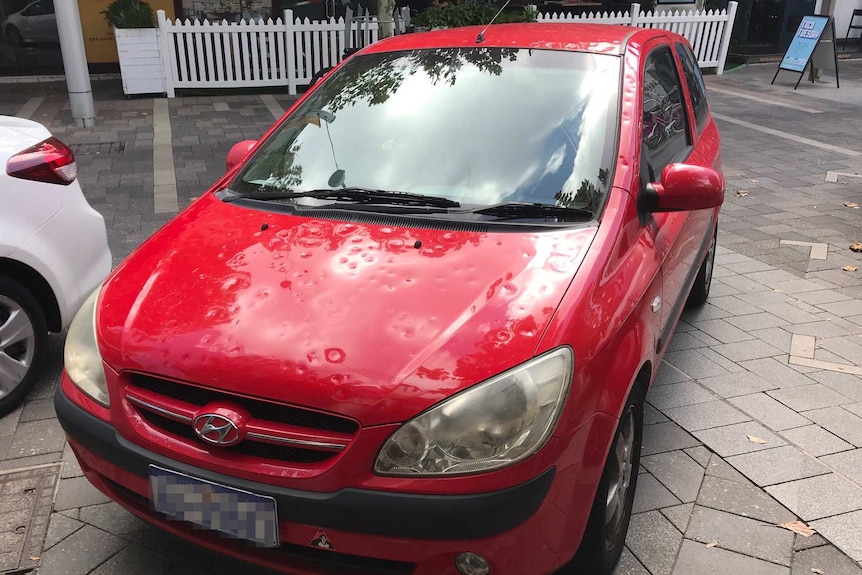 A red car with lots of hail dents in it parked on a road.