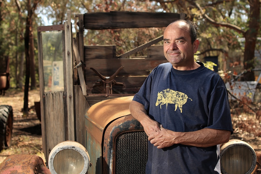 A man in a navy blue t-shirt stands in front of a rusted, old car