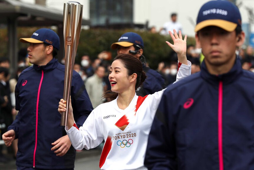An official Tokyo 2020 ambassador waves to a crowd during an Olympic torch relay rehearsal flanked by security guards